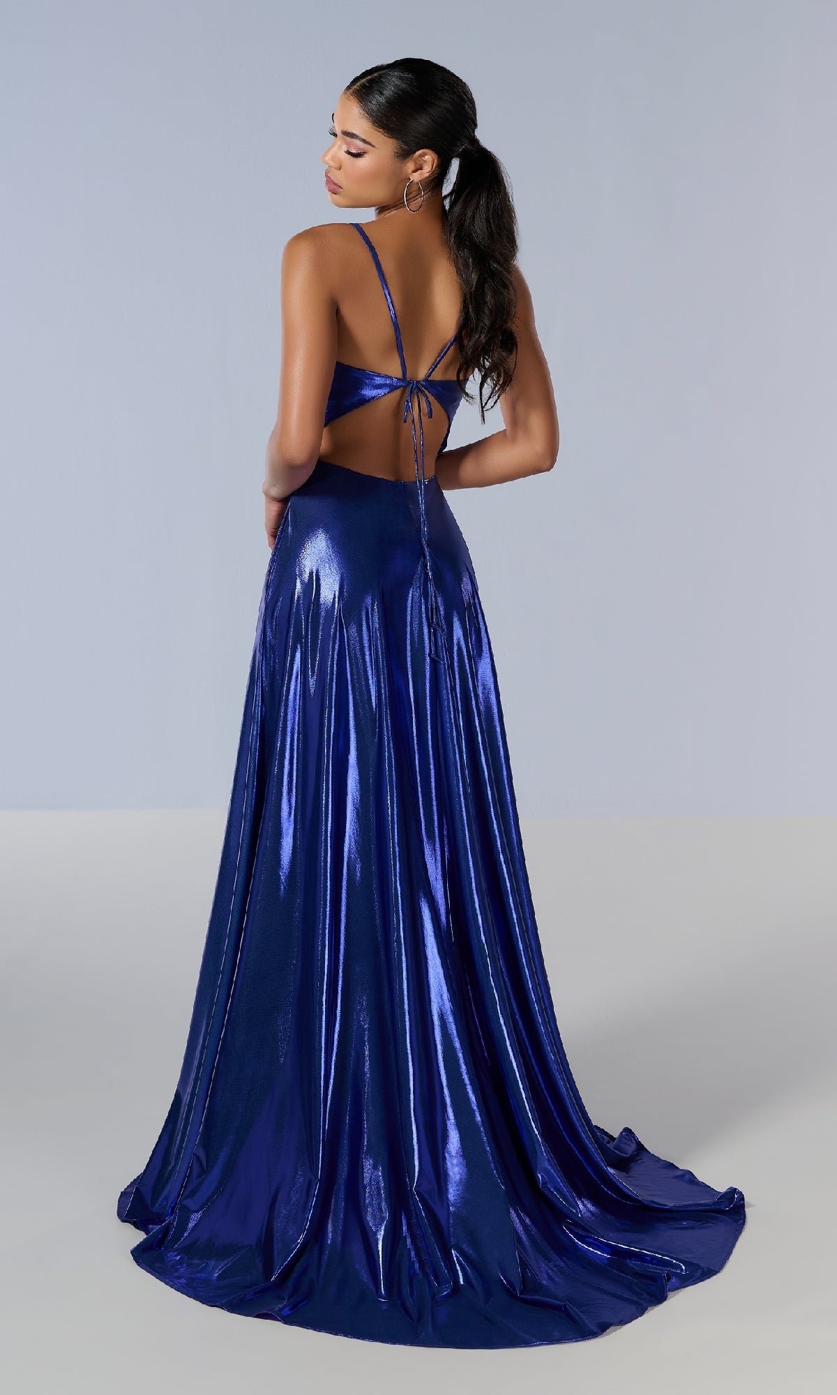 A woman stands with her back to the camera, wearing the Tiffany 16179 long prom dress—a shiny, metallic blue A-line gown with an open back and a flowing skirt.
